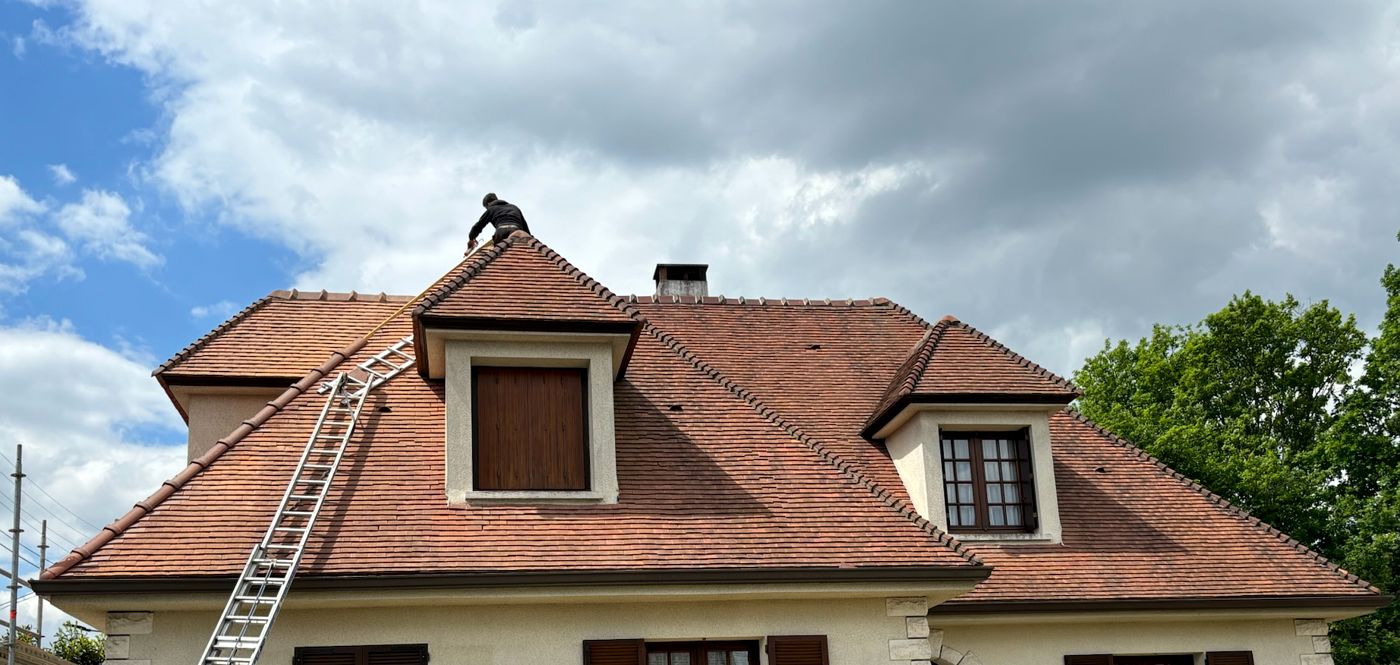 Faîtage et lucarnes d'une toiture en tuiles plates à Palaiseau, un couvreur au sommet du toit
